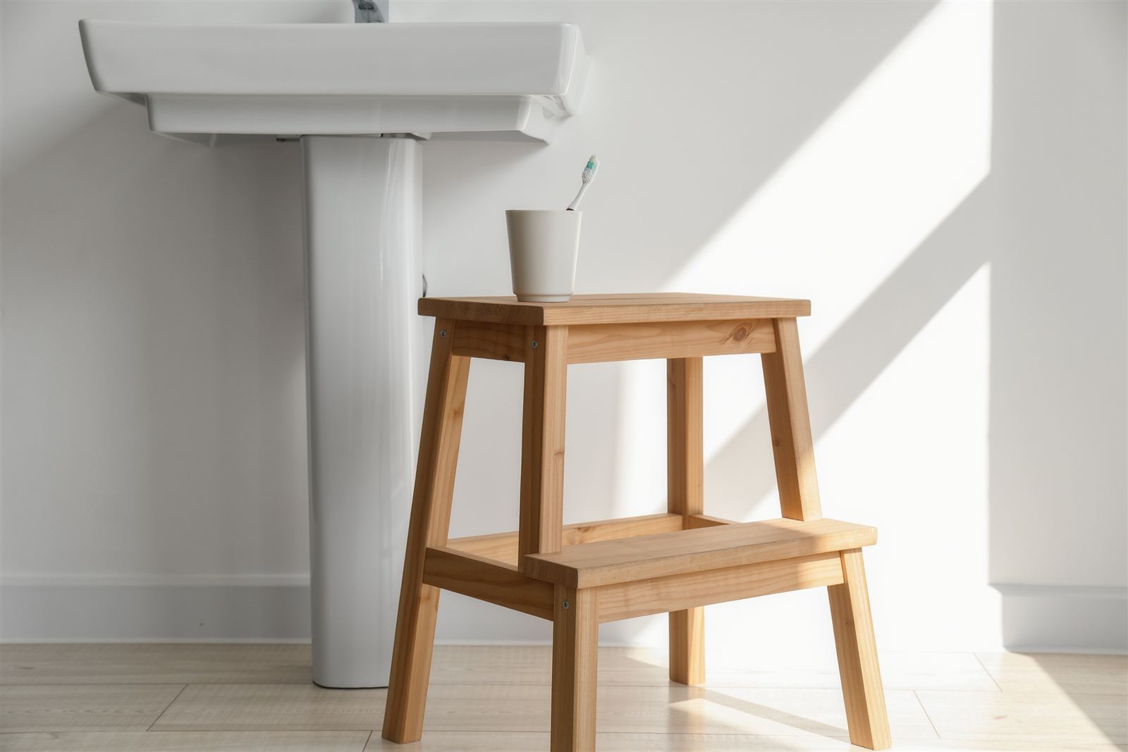 A wooden step stool beneath a white pedestal sink with a toothbrush in a cup